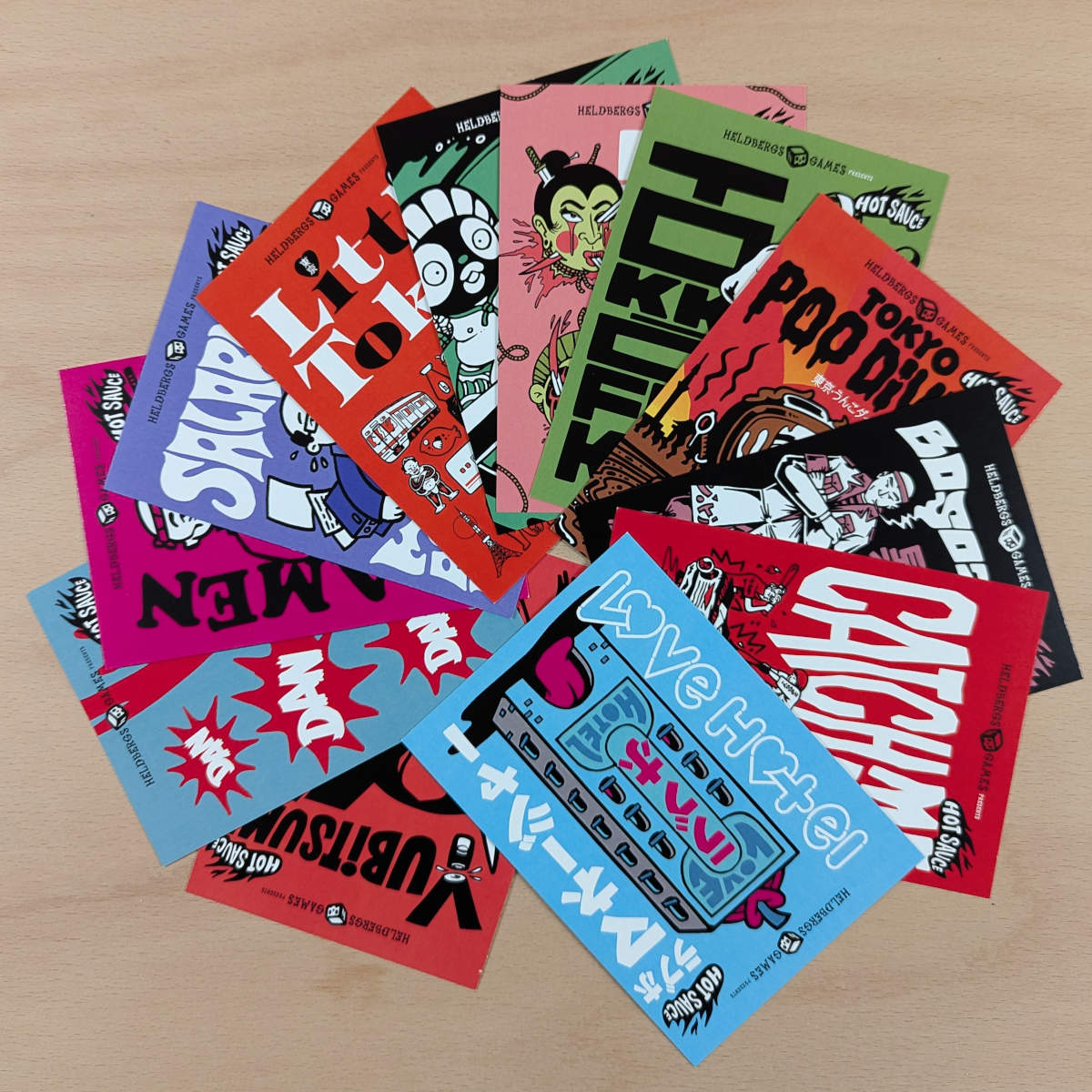 Sammlung von Heldbergs Games Postkarten, fächerförmig angeordnet, mit Designs wie Namakubi, Tokyo Poo Divers, Catch the Wave. Bunte Farben, Pop-Art-Stil mit japanischen Einflüssen.